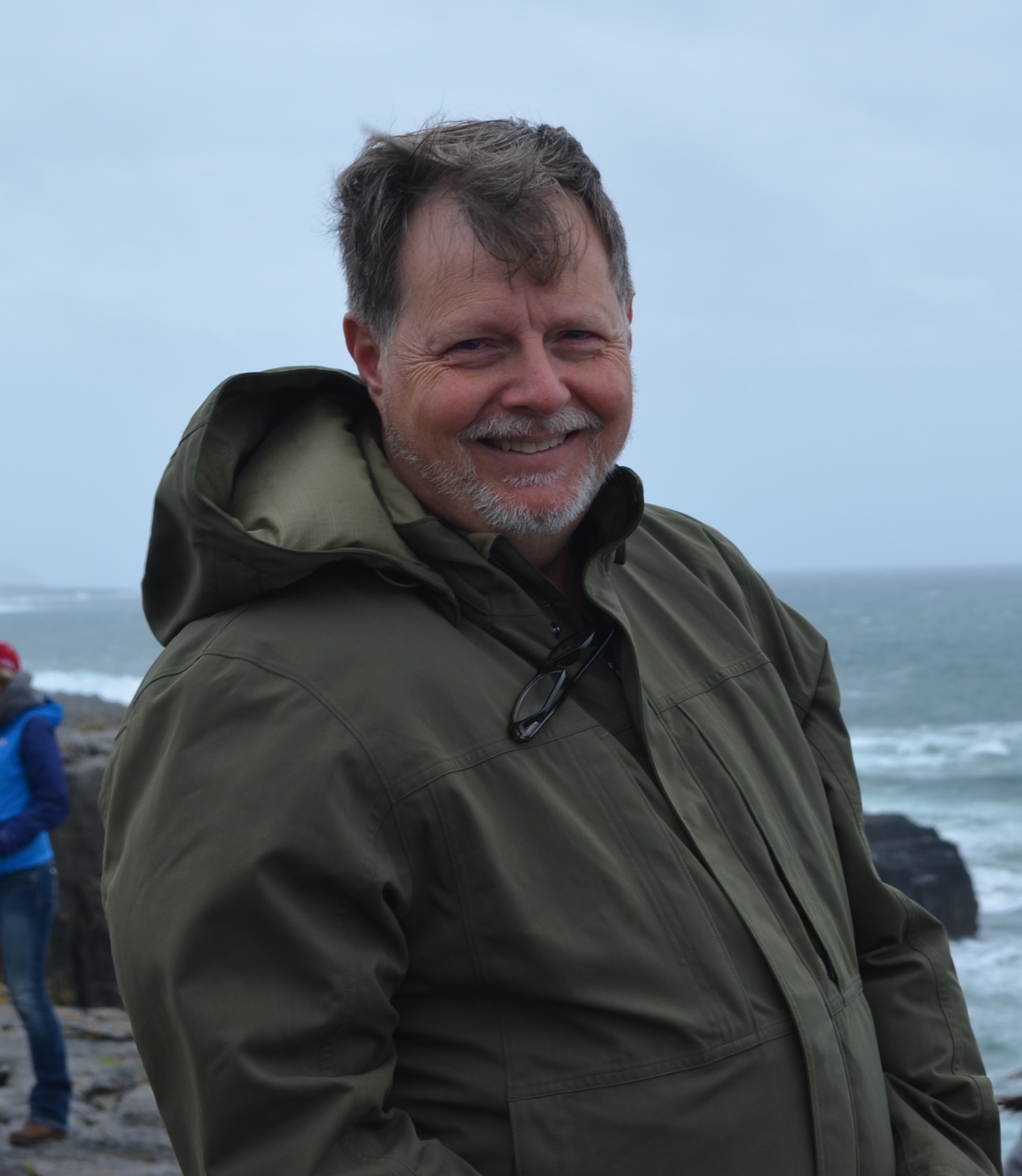 Smiling man in a green jacket standing on a rocky shoreline with ocean waves in the background.