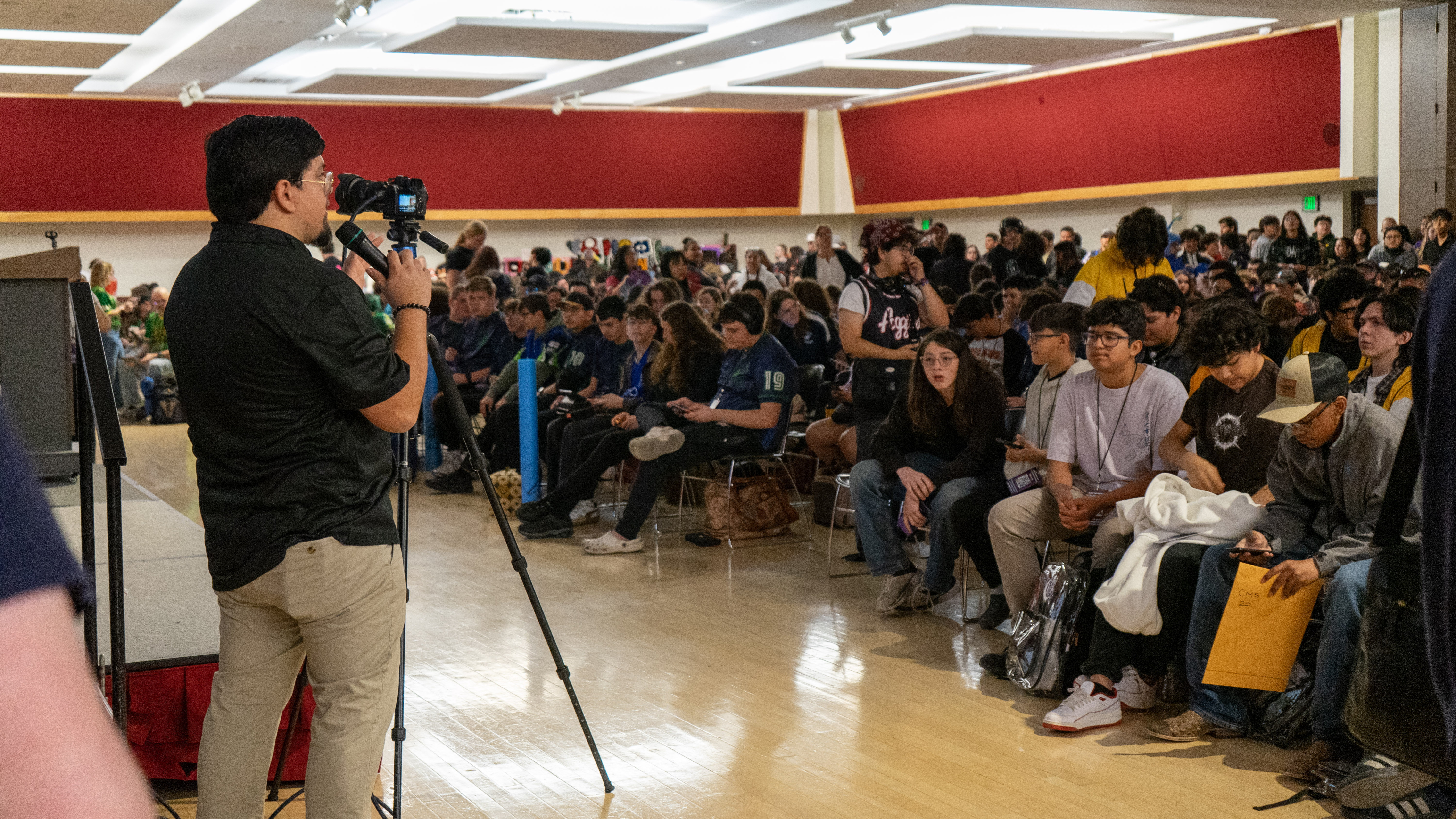 Speaker addresses a large seated audience in a school auditorium during an event.