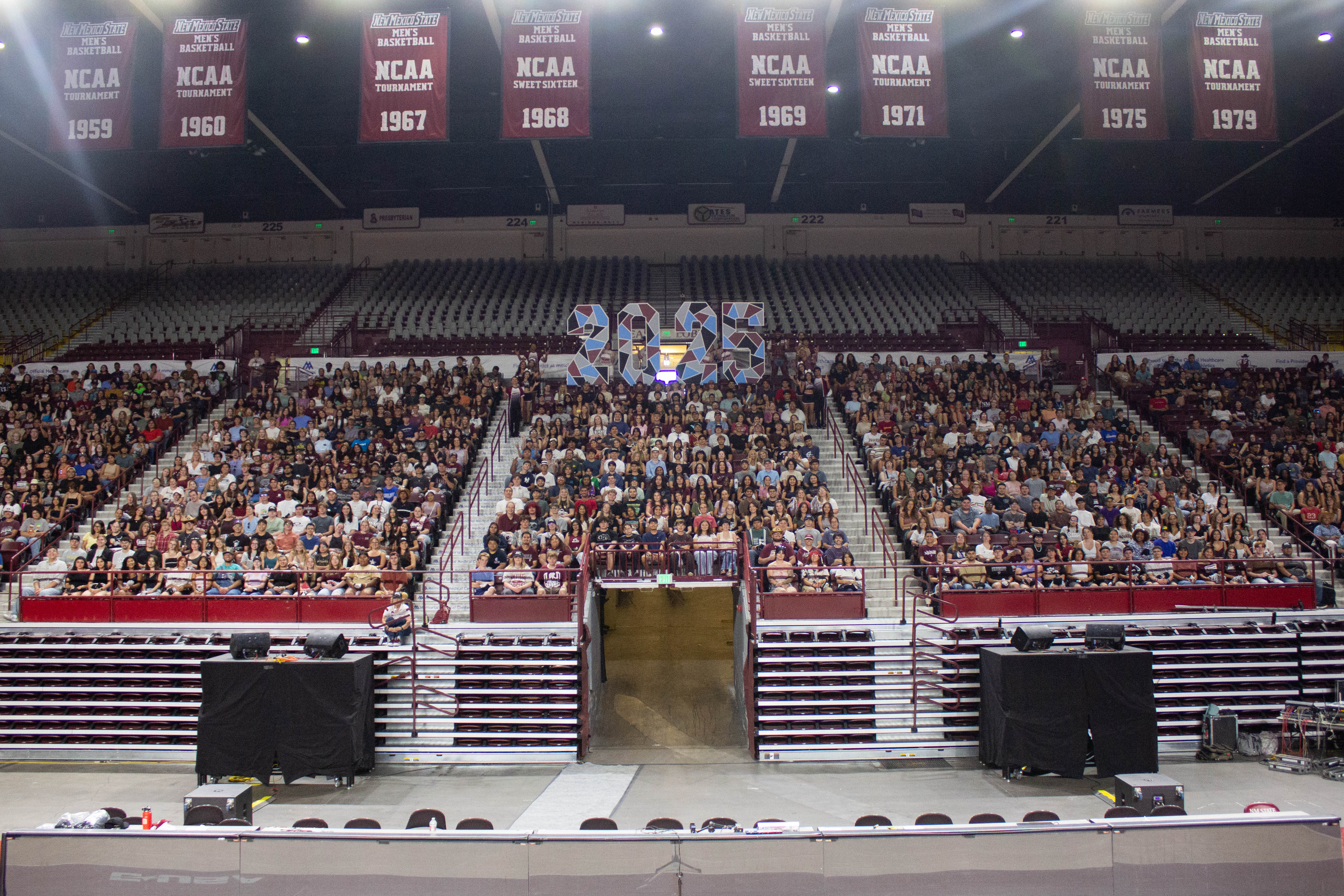 A large audience seated in a sports arena, with banners hanging from the ceiling and the stage visible in the foreground.